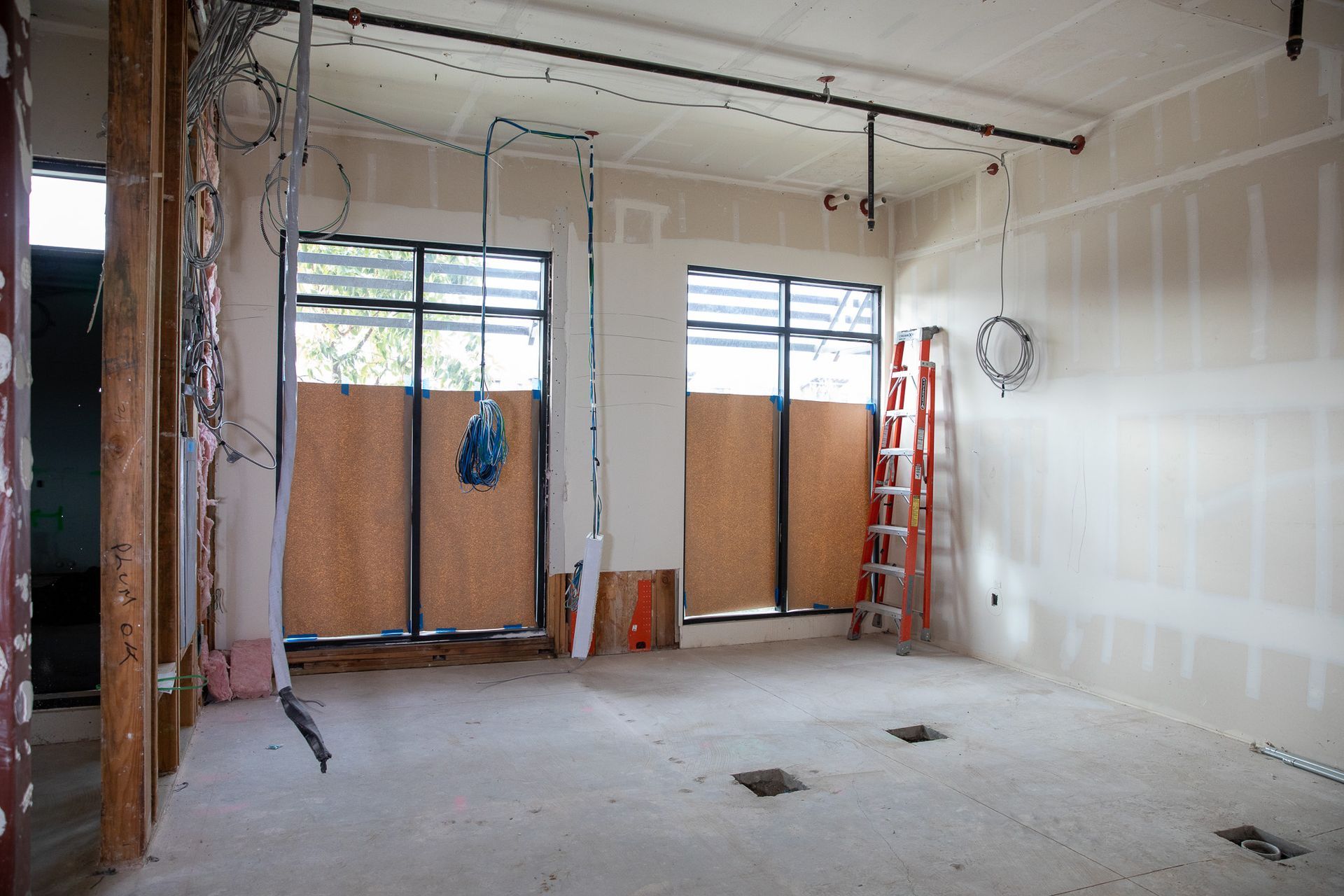 Interior of an unfinished room with exposed drywall, boarded windows, hanging cords, and a ladder.