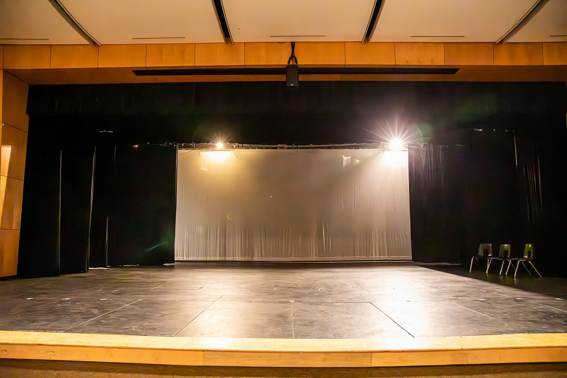 Empty theater stage with dark curtains, a lit backdrop, and a few chairs on the right