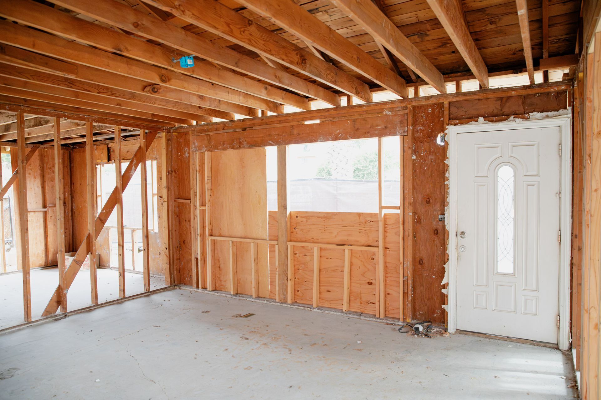 Exposed wooden framing in an unfinished room with a concrete floor and a white exterior door.