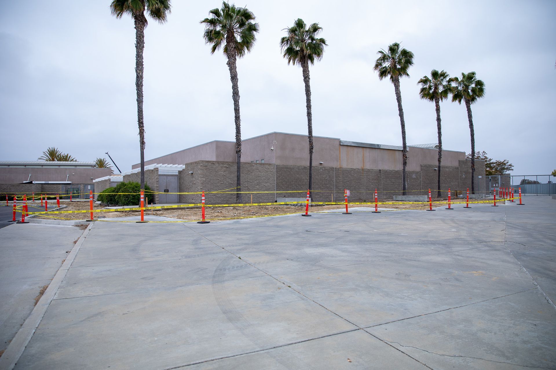 Empty parking lot beside a large building with palm trees and orange safety cones under a cloudy sky
