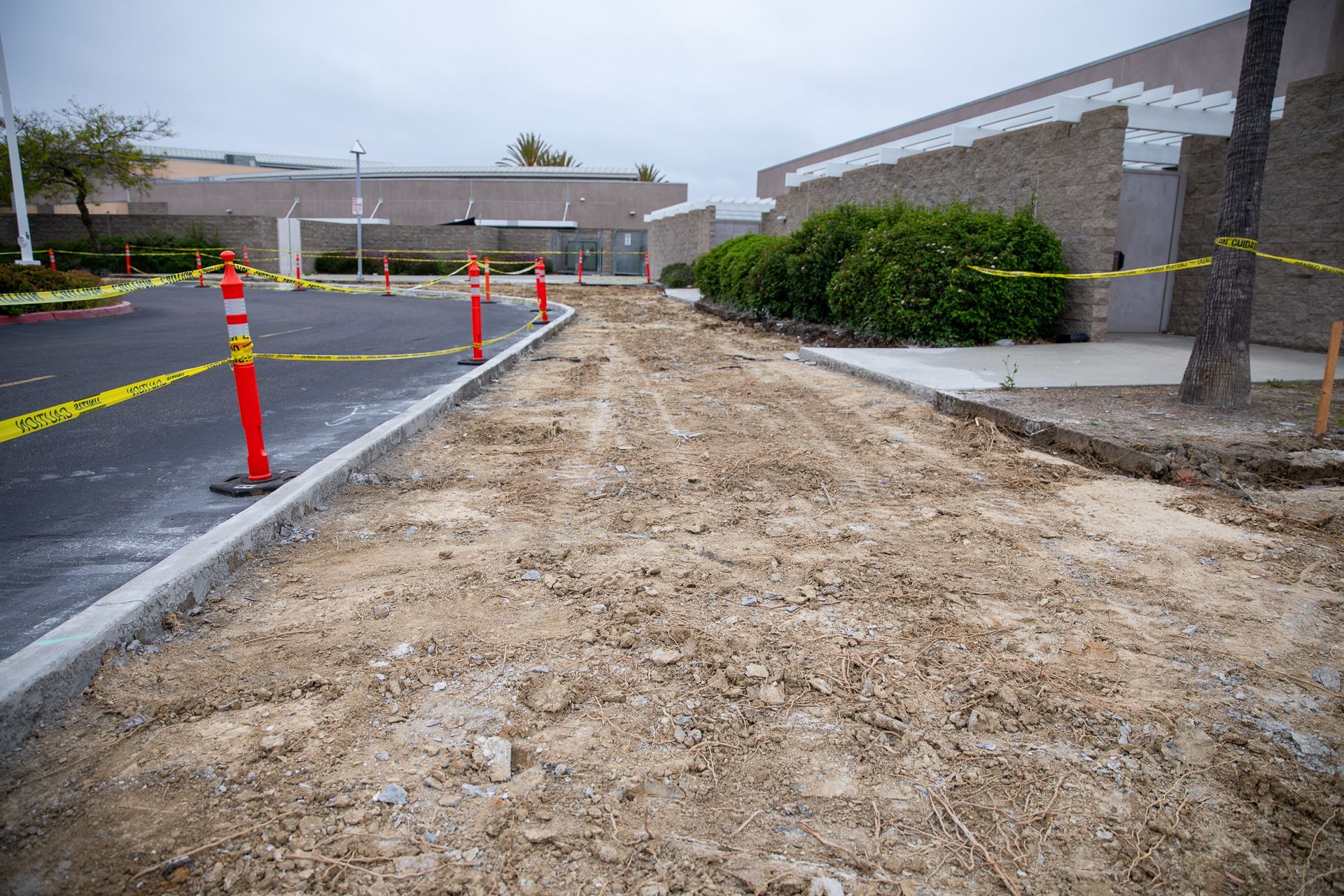Construction area beside a building with orange cones, caution tape, and a dirt walkway beside pavement.