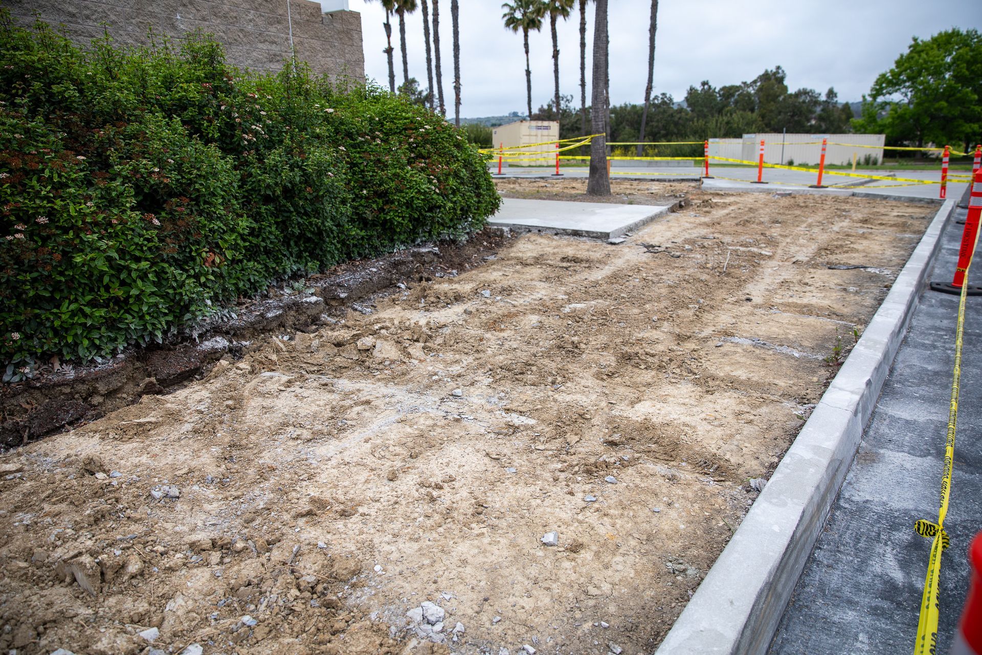 Freshly graded dirt lot beside a sidewalk, with orange cones and green hedges near palm trees