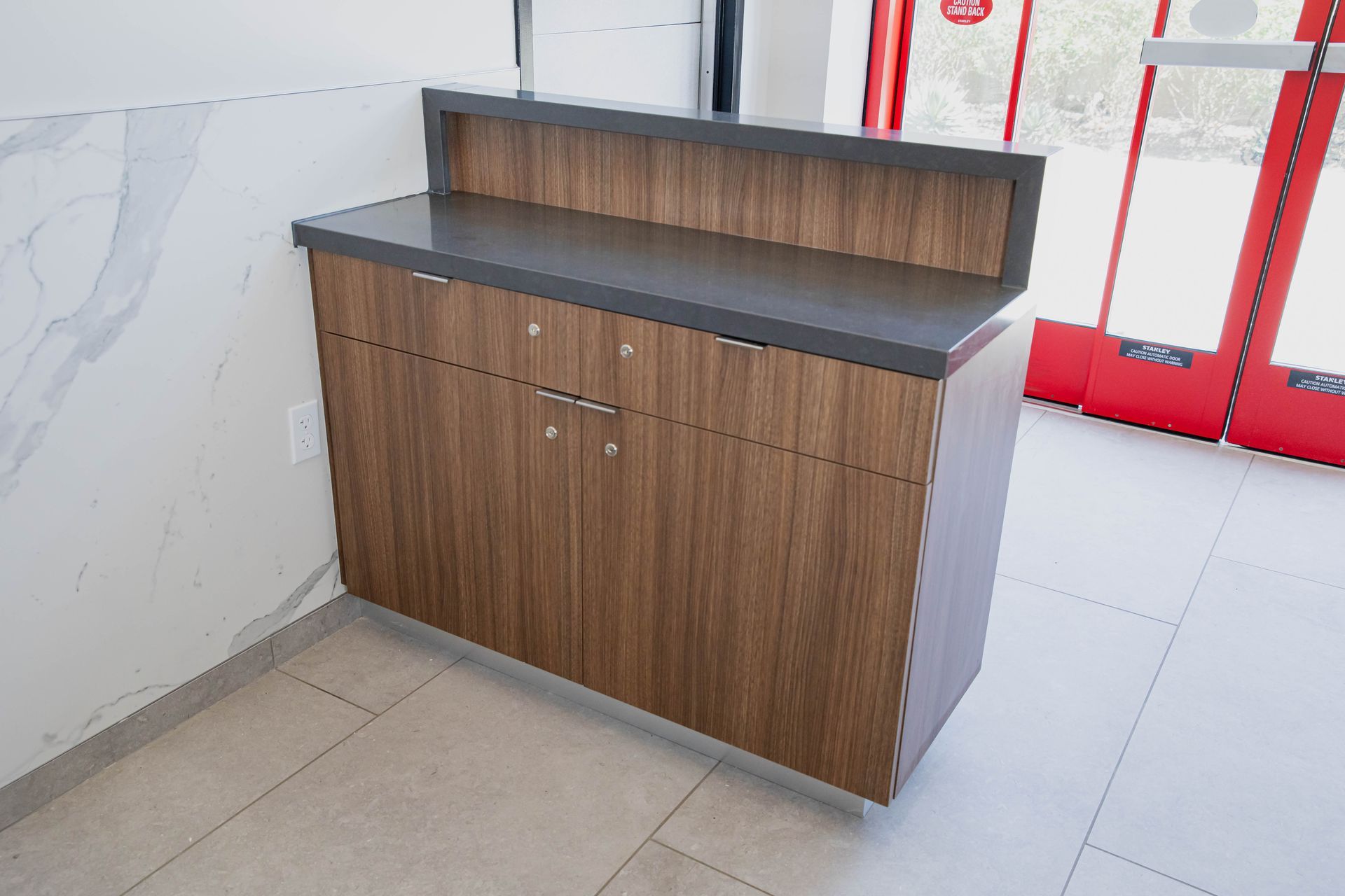 Wooden cabinet with black countertop and backsplash in a bright tiled room, near a red-framed glass door