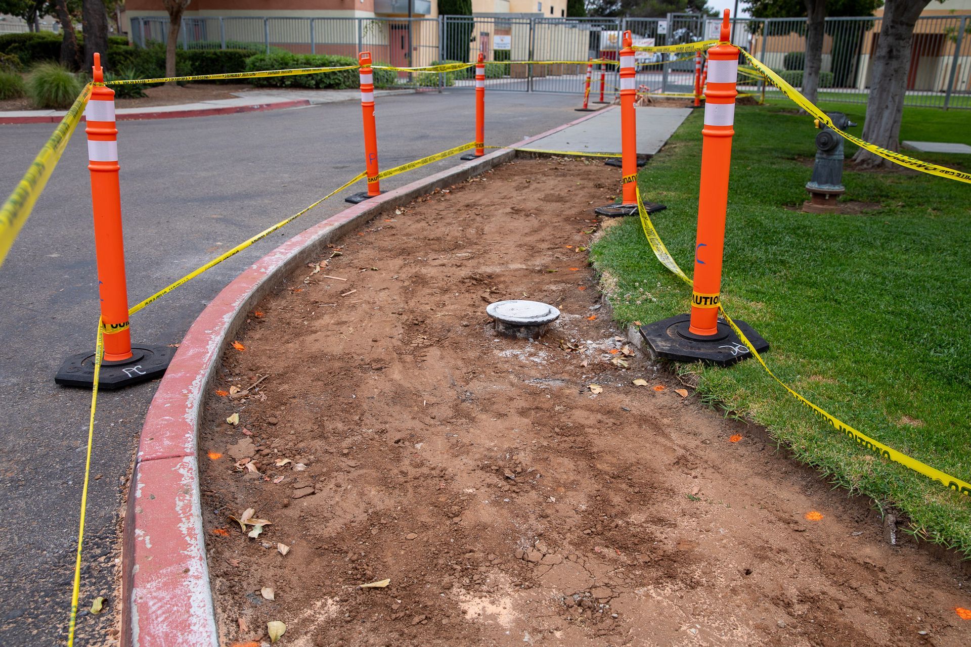 Orange construction cones and caution tape marking a dirt path beside a paved road and grass.