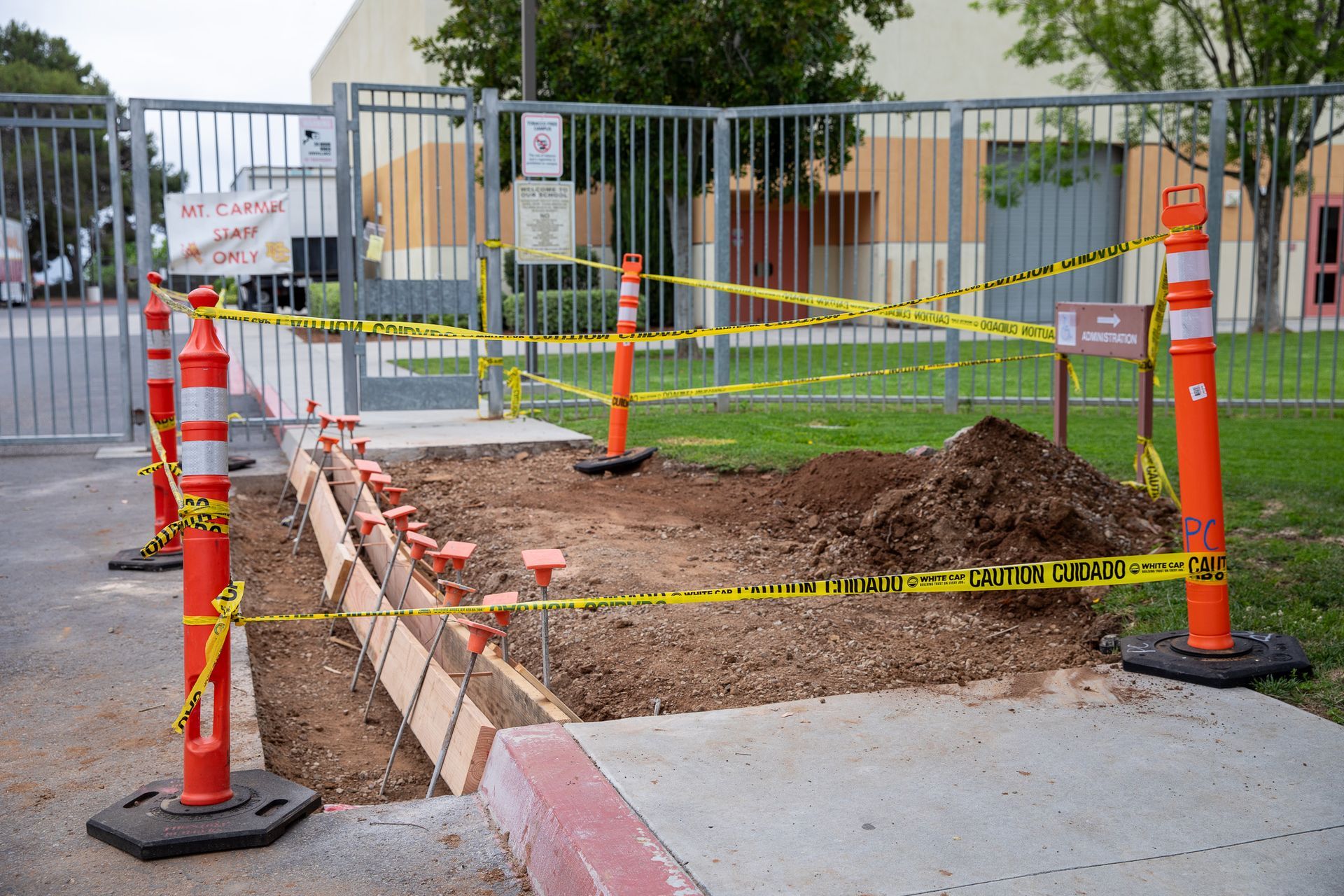 Cordoned-off sidewalk excavation with orange cones, caution tape, and a dirt-filled trench near a gate