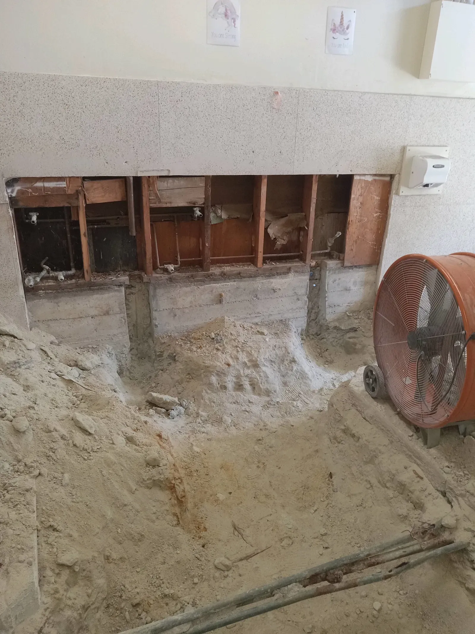 A fan is sitting on the floor next to a hole in the wall.