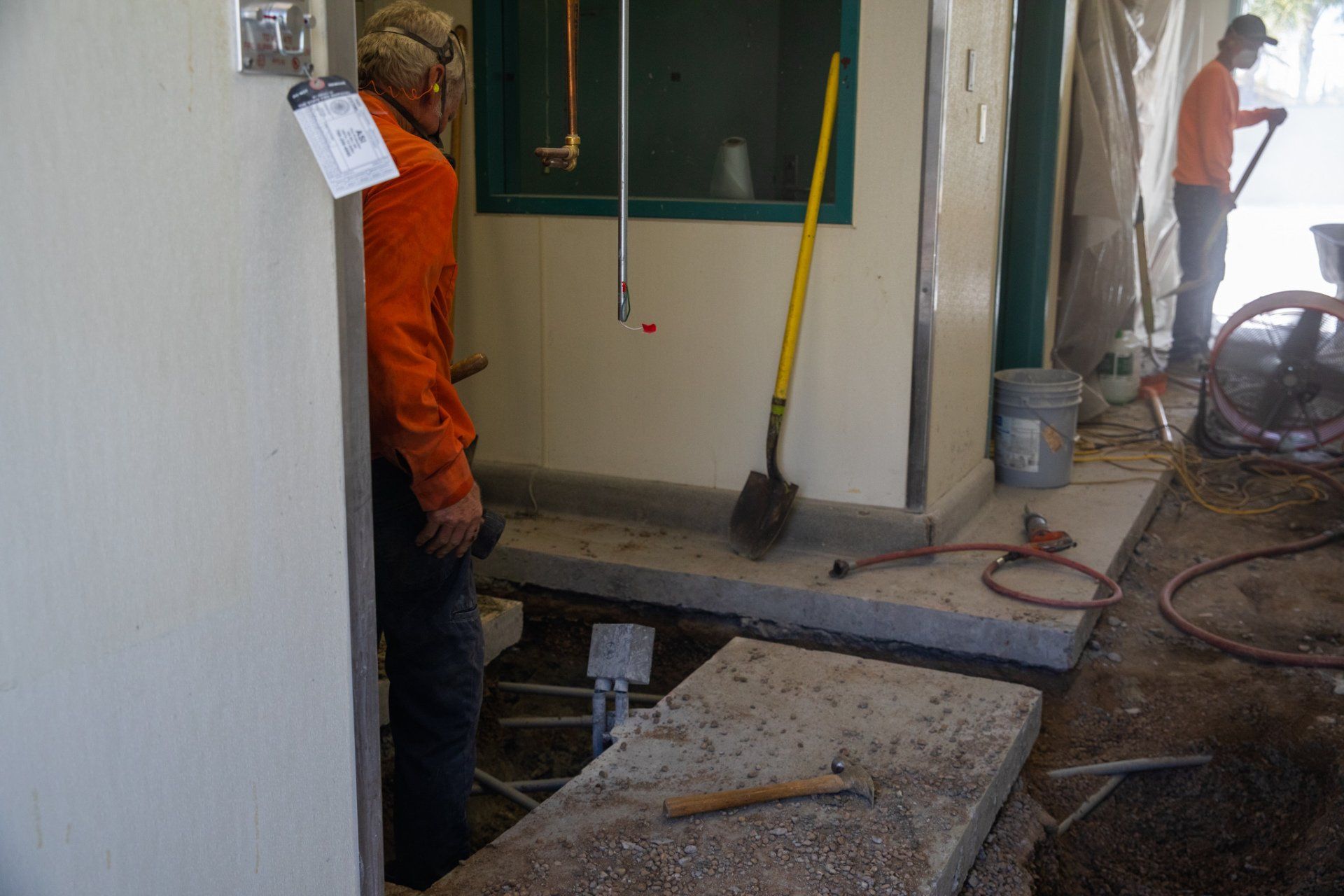 A man in an orange jacket is standing in a room with a shovel.
