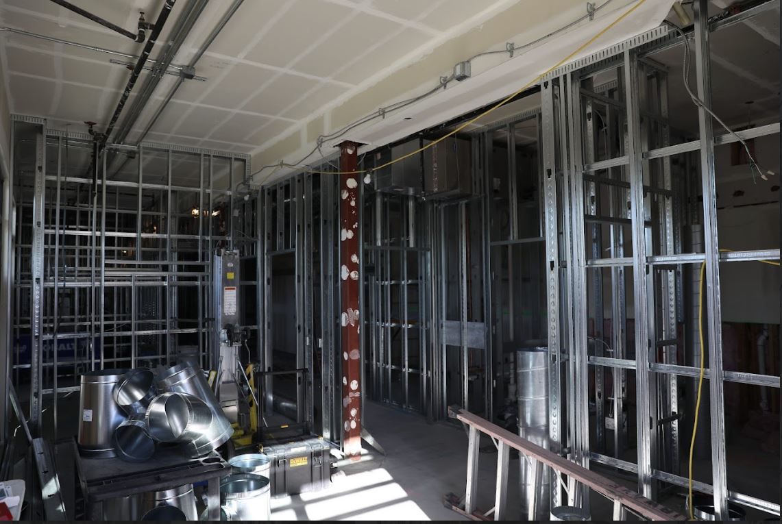 Interior of a room under construction with exposed metal framing and wiring, white ceiling, and sunlight on the floor