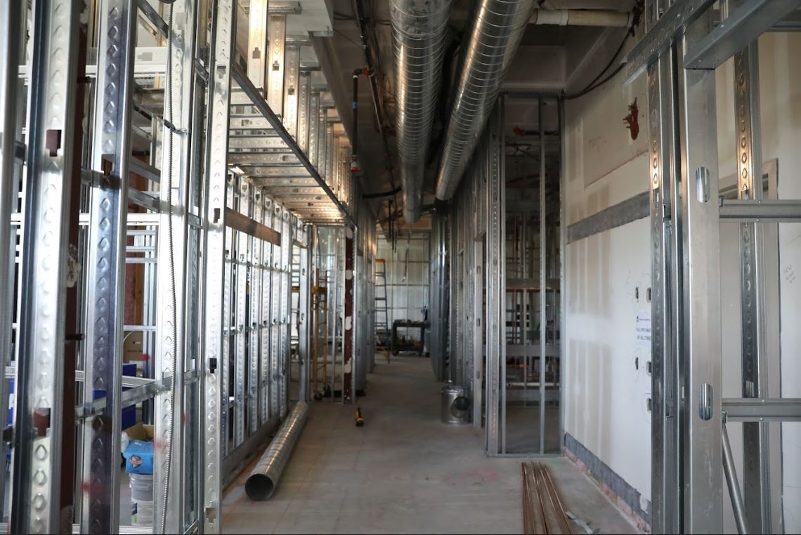 Interior of a building under construction with exposed metal studs and unfinished walls along a narrow hallway.