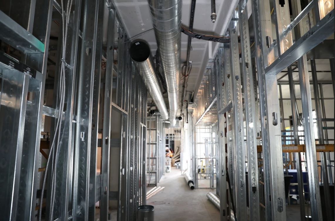 Metal-framed interior under construction with exposed ducts, pipes, and unfinished walls along a narrow corridor