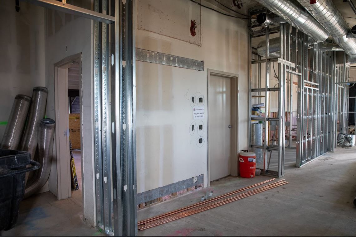 Unfinished interior with metal wall framing, utility panels, ladders, and construction materials along a corridor.
