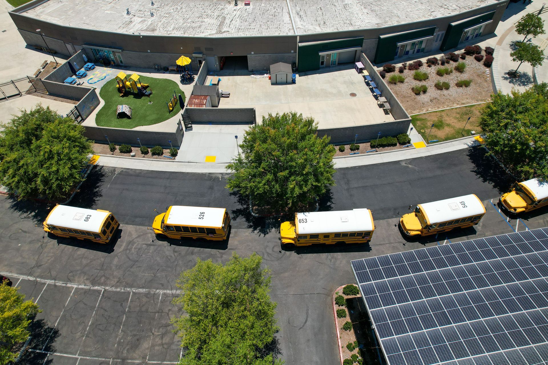Aerial view of several yellow-and-white buses parked along a curved road beside a building and solar panels.