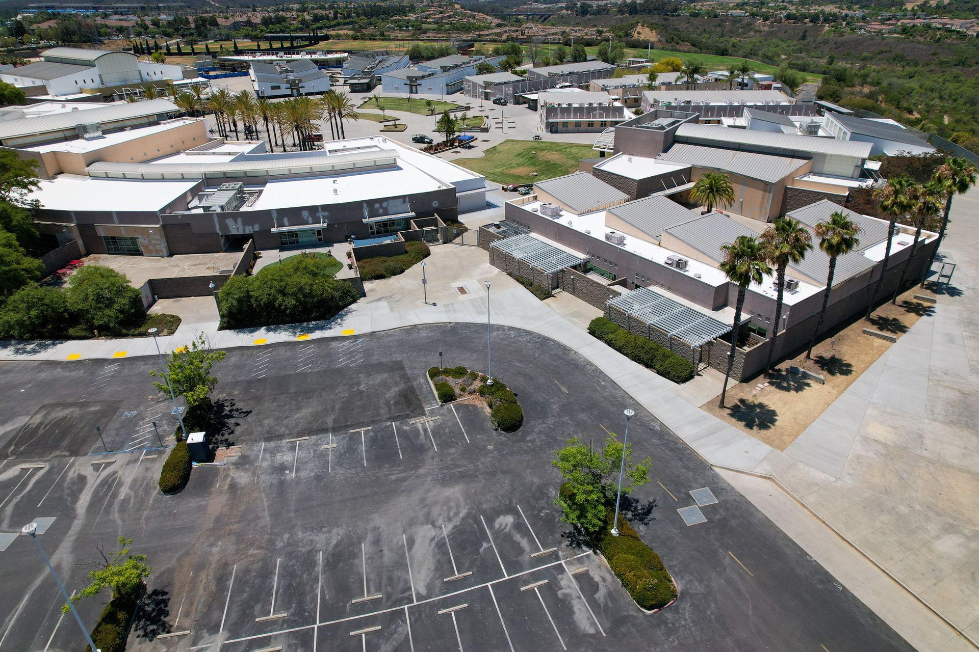 Aerial view of a school campus with parking lots, buildings, trees, and a curved road under clear skies