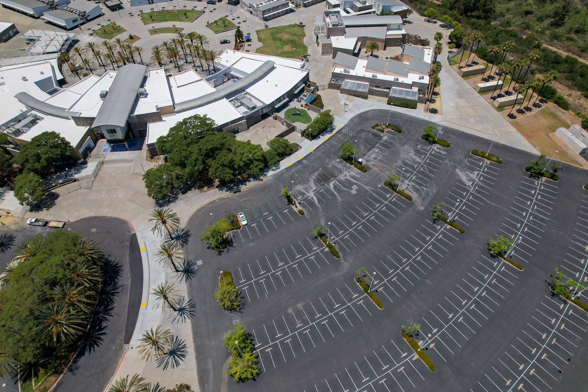 Aerial view of a large mostly empty parking lot beside buildings and trees on a sunny day