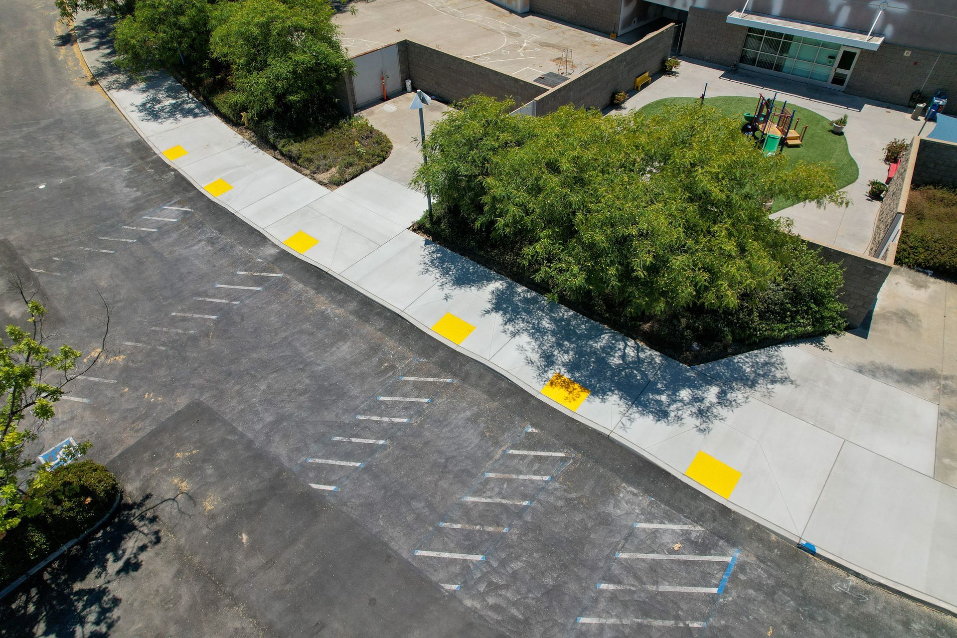 Aerial view of a sidewalk beside a parking lot and playground, with yellow curb markers and trees.