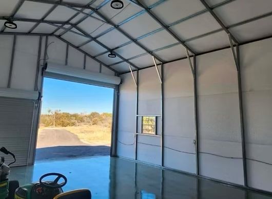 Inside view of a large, empty metal building with a roll-up door open to the outdoors, showing a sunny landscape.
