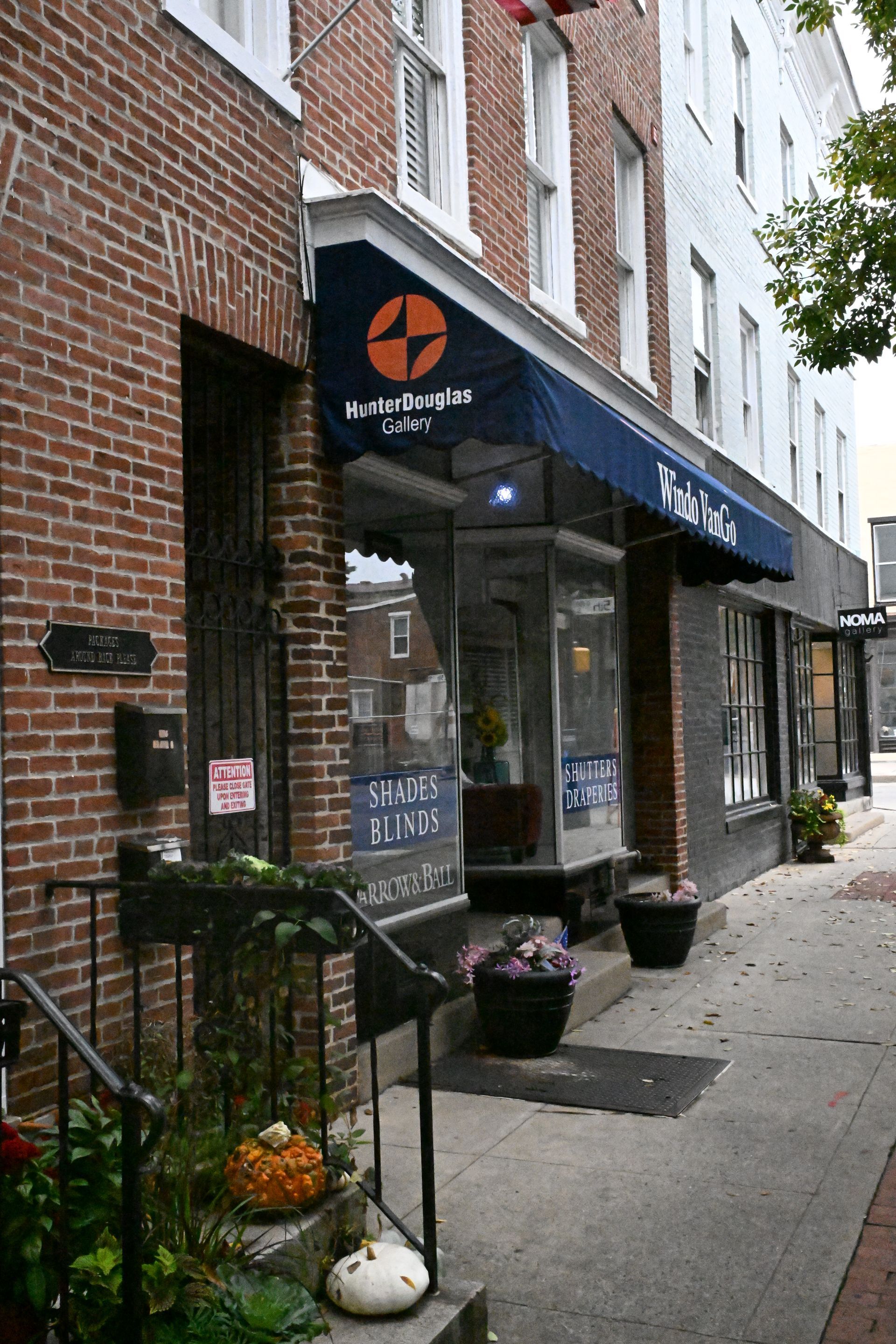 A sidewalk view of brick storefronts with a blue awning, potted plants, and seasonal gourds on the front steps.