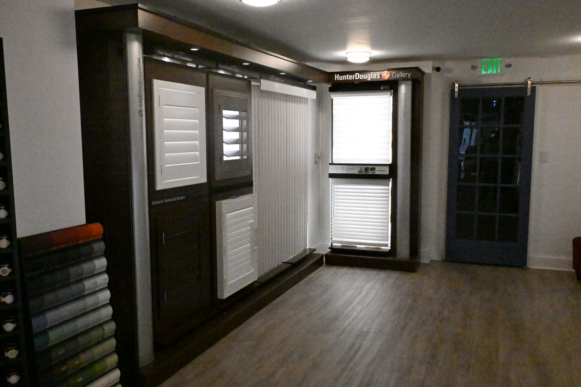 A showroom display featuring various window blinds and shutters, with fabric samples and a dark-framed glass door nearby.