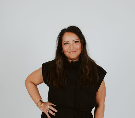 Smiling person in a black sleeveless top, standing with hand on hip against a plain light background