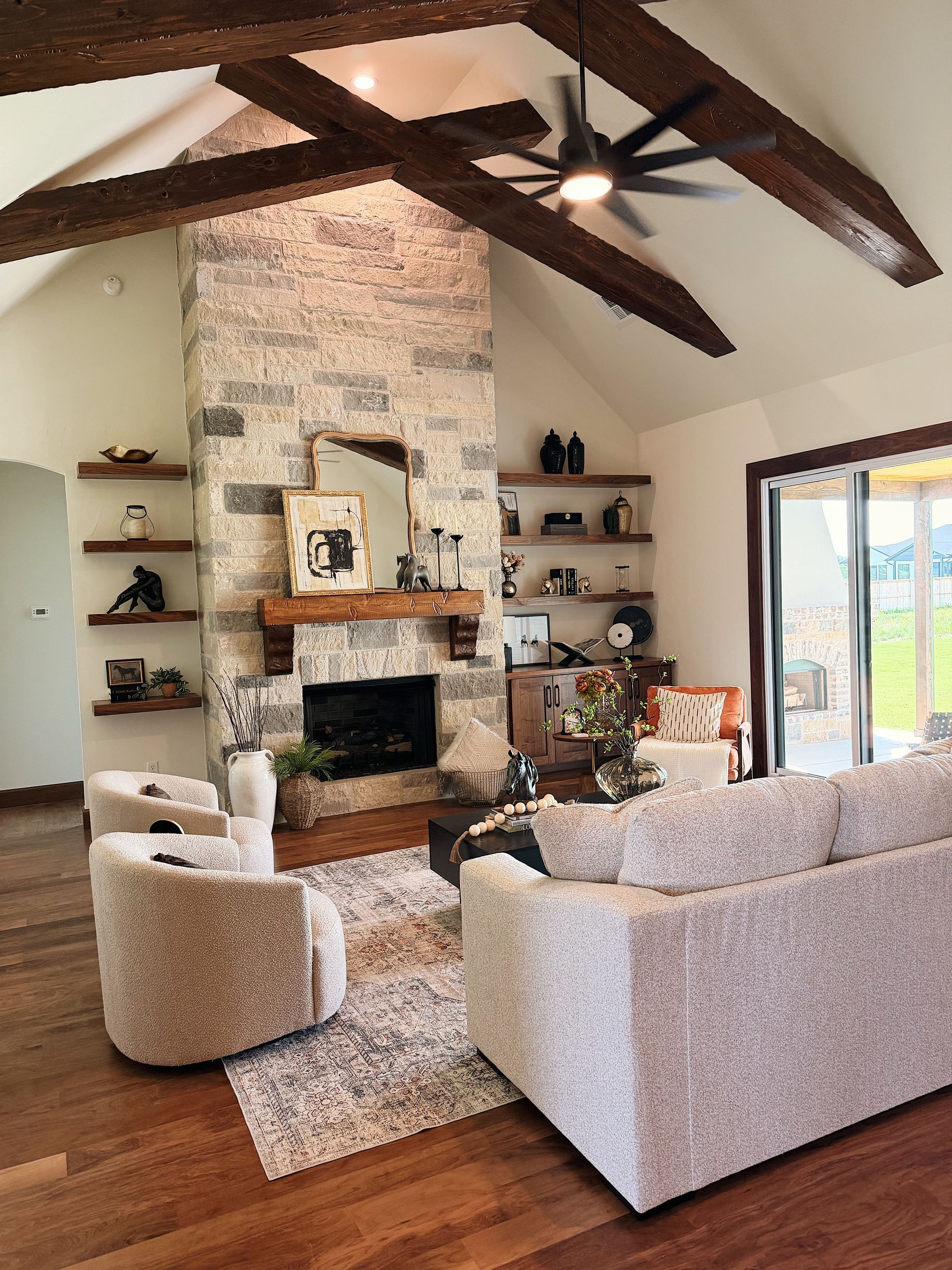 Bright living room with stone fireplace, exposed wood beams, beige sofas, and open patio doors