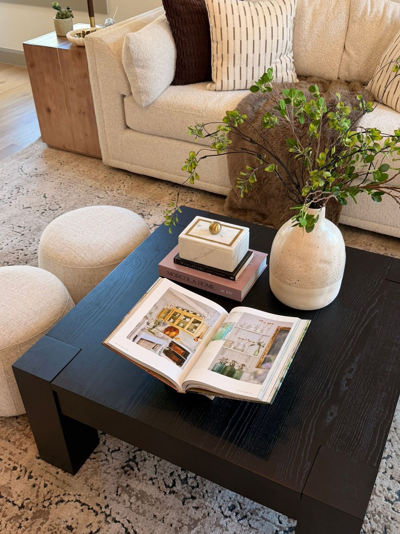 Cozy living room with black coffee table, open book, vase of greenery, beige sofa, and pouf.