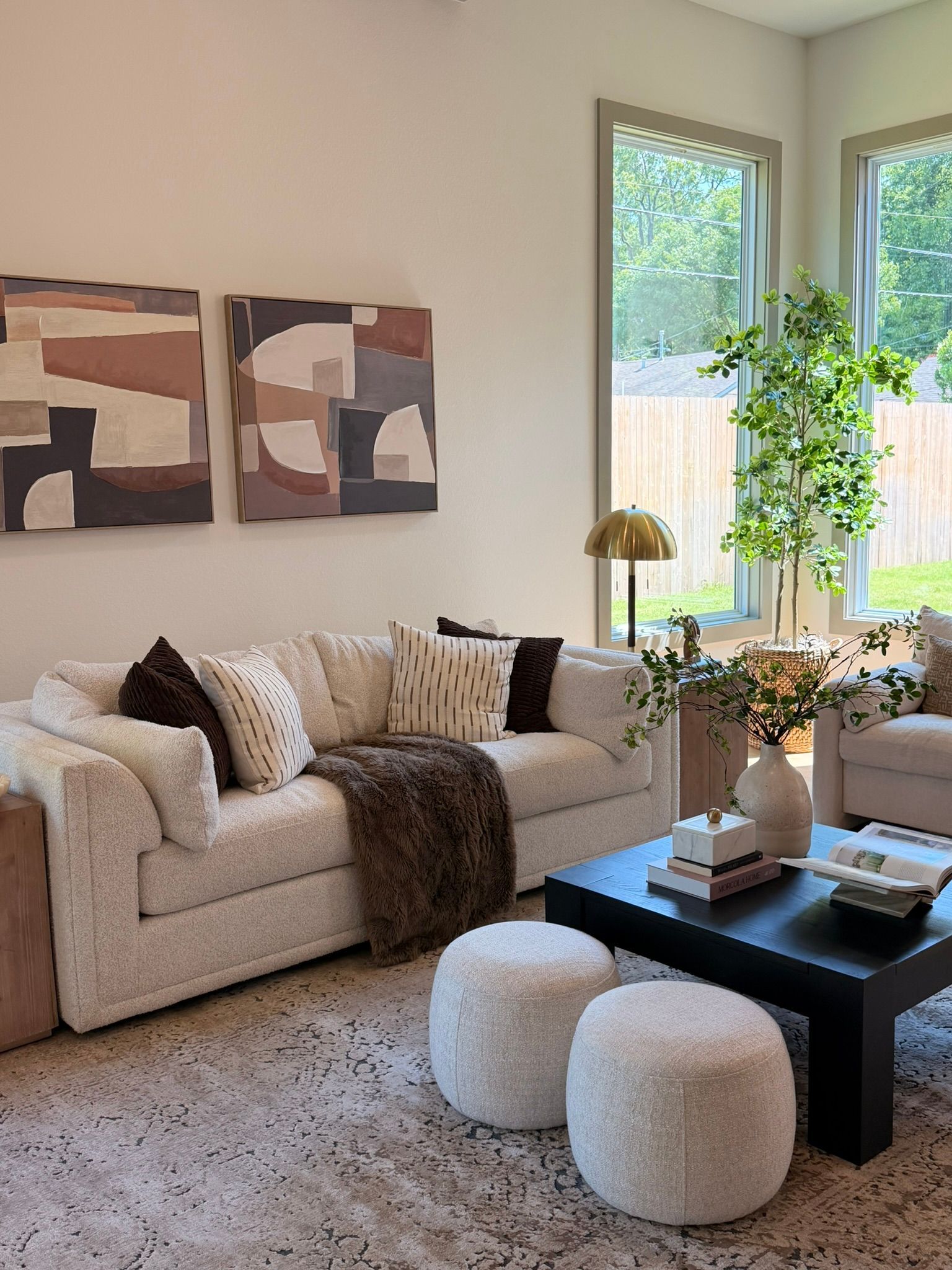 Cozy living room with beige sofa, black coffee table, poufs, wall art, and tall windows with greenery.