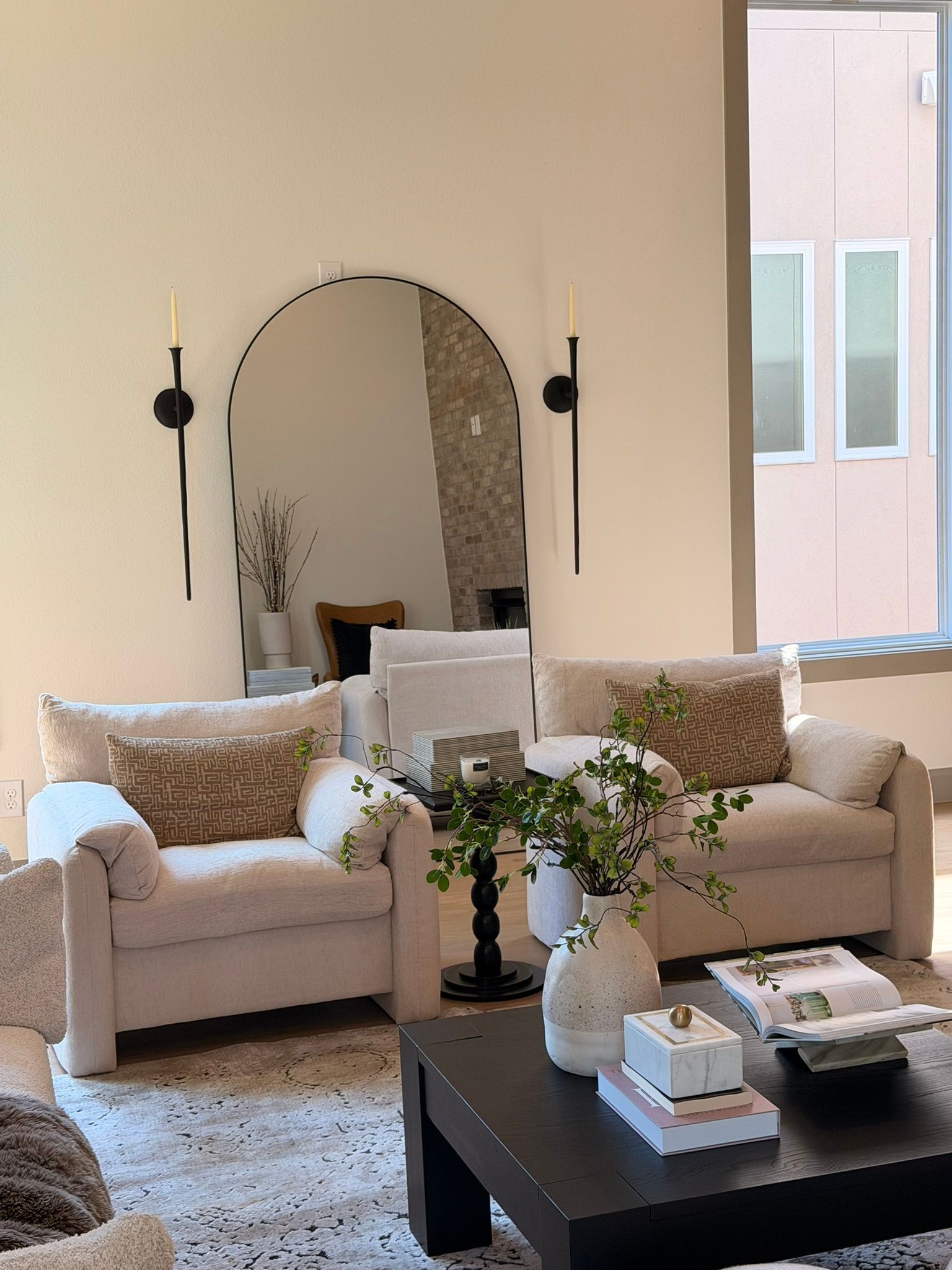 Cozy beige living room with two armchairs, a sofa, arched mirror, and black coffee table with flowers.