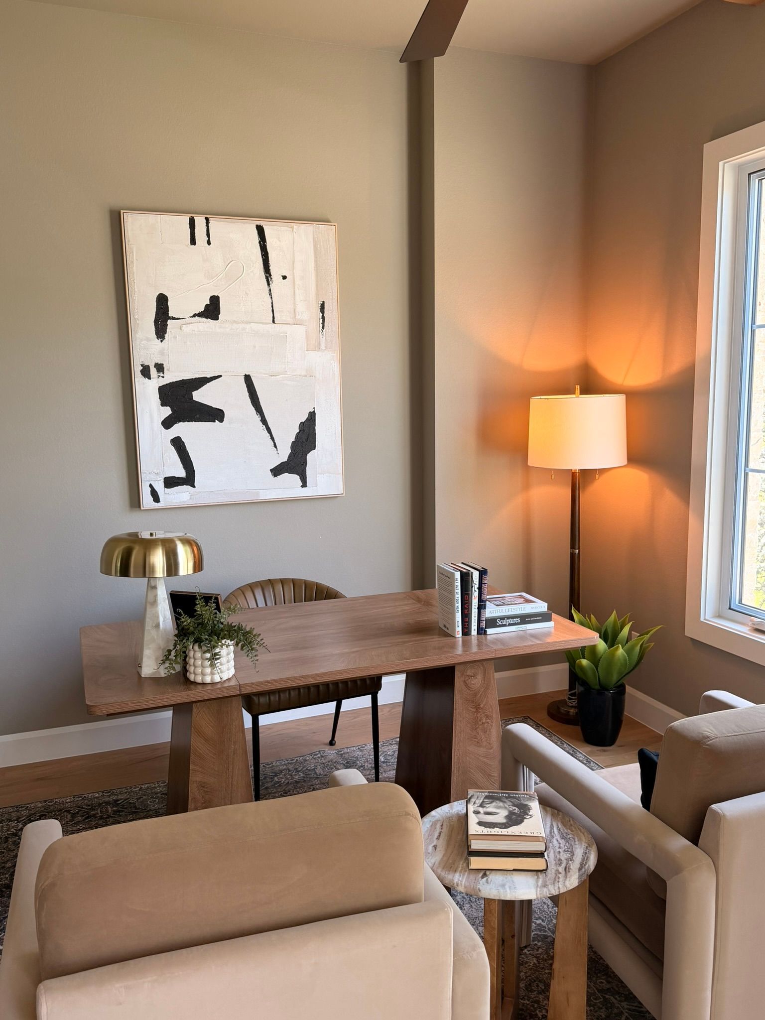 Cozy living room with beige chairs, wooden table, floor lamp, abstract wall art, and a potted plant.