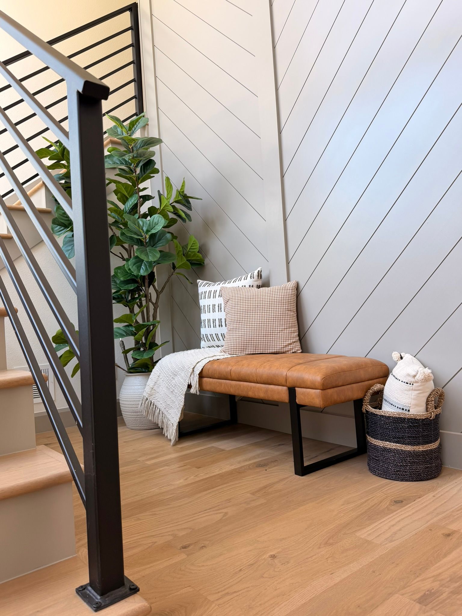 Modern staircase nook with tan bench, patterned pillows, throw blanket, plant, and black metal railing