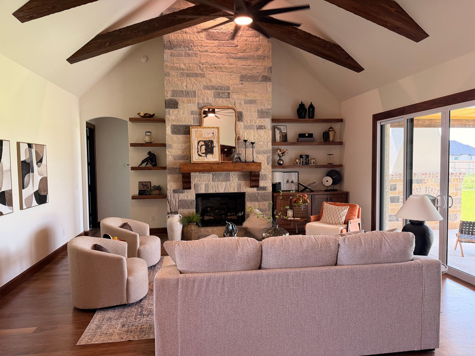 Cozy living room with brick fireplace, exposed beams, beige sofa, armchairs, shelves, and sliding glass door