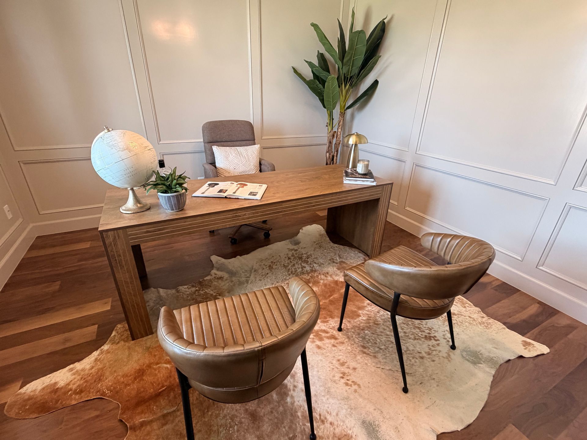 Modern office with wooden desk, two brown chairs, rug, globe lamp, and tall plant in a bright room