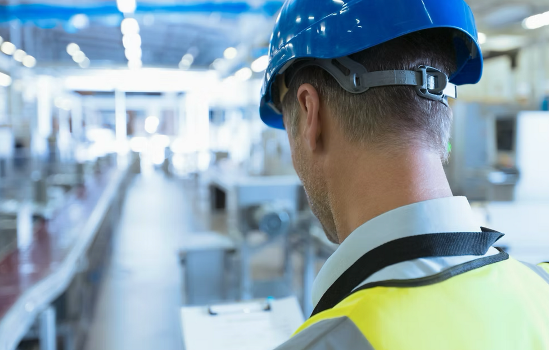 A man wearing a hard hat and safety vest is standing in a factory and reading a clipboard