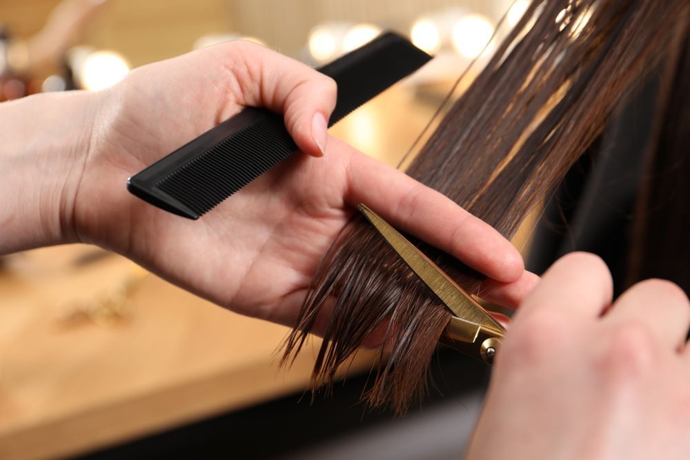 Hands holding comb and scissors, cutting wet brown hair in salon.