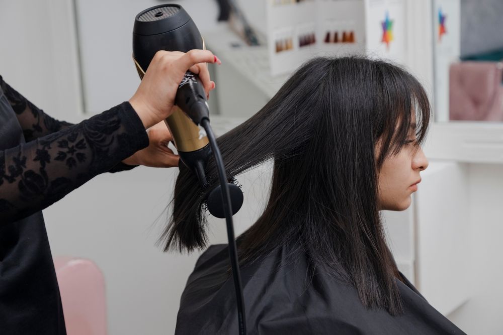 Hairdresser using a hair dryer on a client's dark hair in a salon.