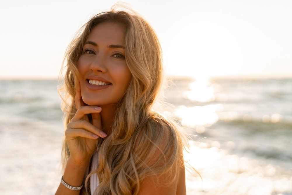 Woman with blonde hair smiles at the camera on a beach with ocean and sunlight in the background.
