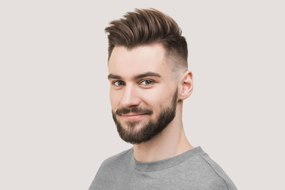 Man with brown hair and beard, wearing a gray shirt, smiling against a neutral background.