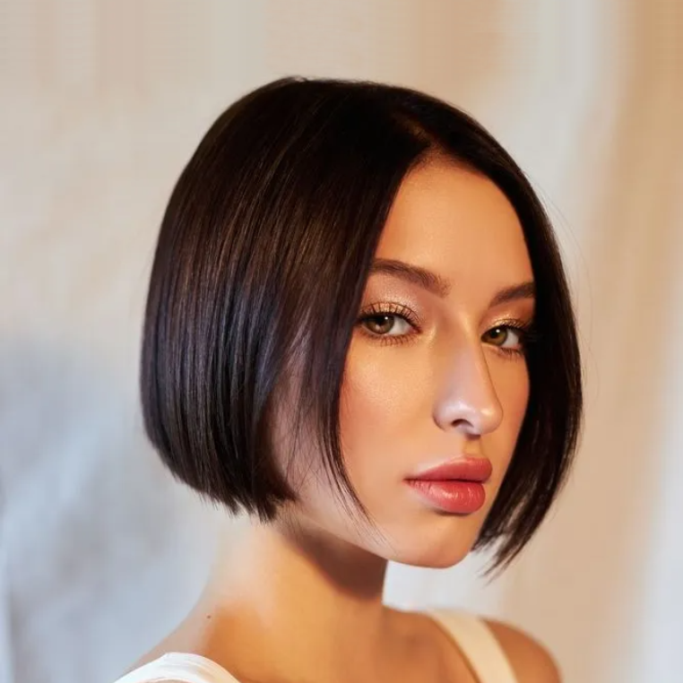 Woman with short dark hair, wearing a white tank top, looking towards the camera.