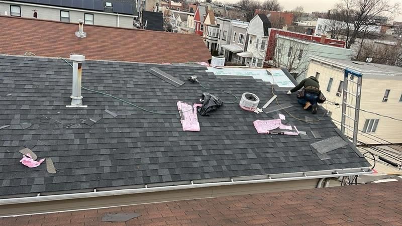 Roofer works on a shingled roof with supplies, residential buildings in the background. Overcast day.