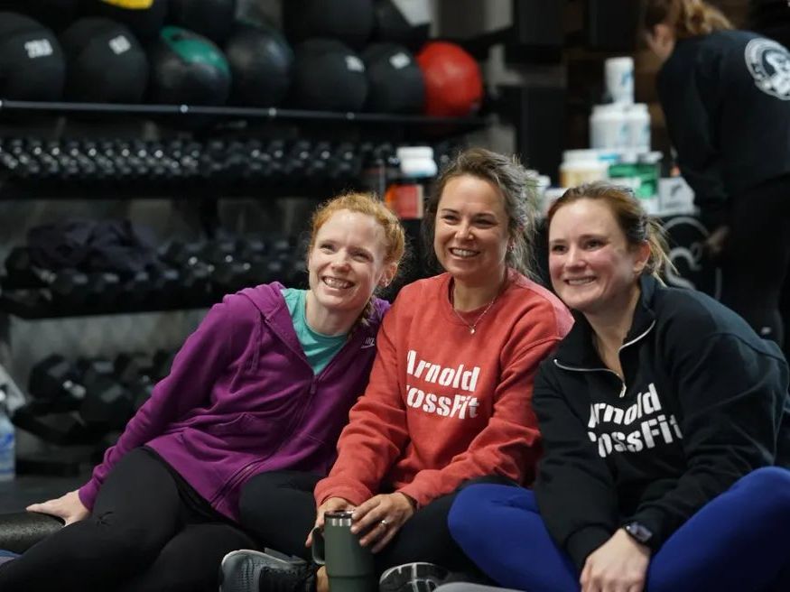 Three smiling women in athletic wear pose together in a gym, with weights and equipment in the background.