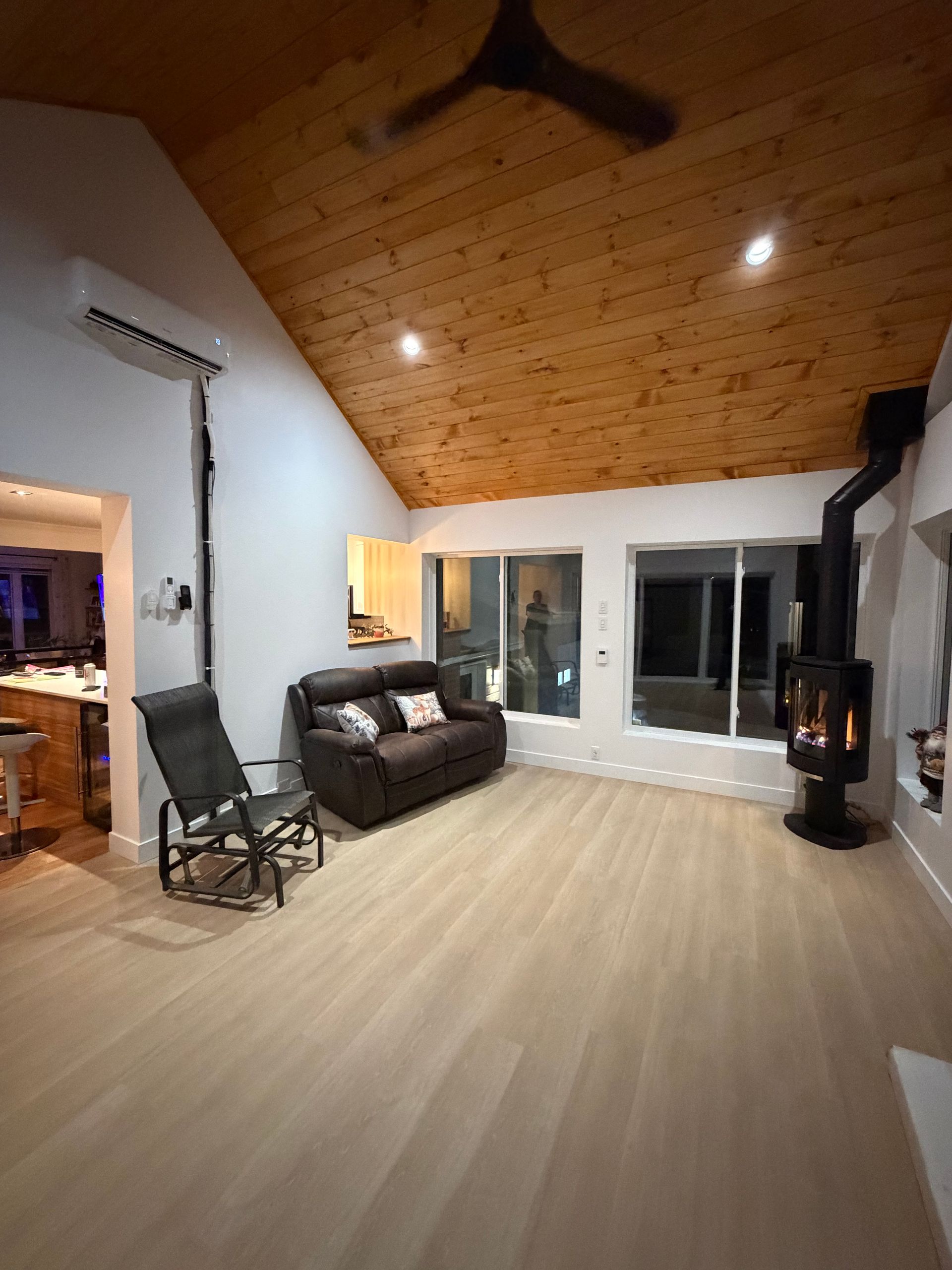 Salon chaleureux avec plafond en bois, fenêtres et poêle à bois. Comprend un canapé deux places, un fauteuil à bascule et un parquet clair.