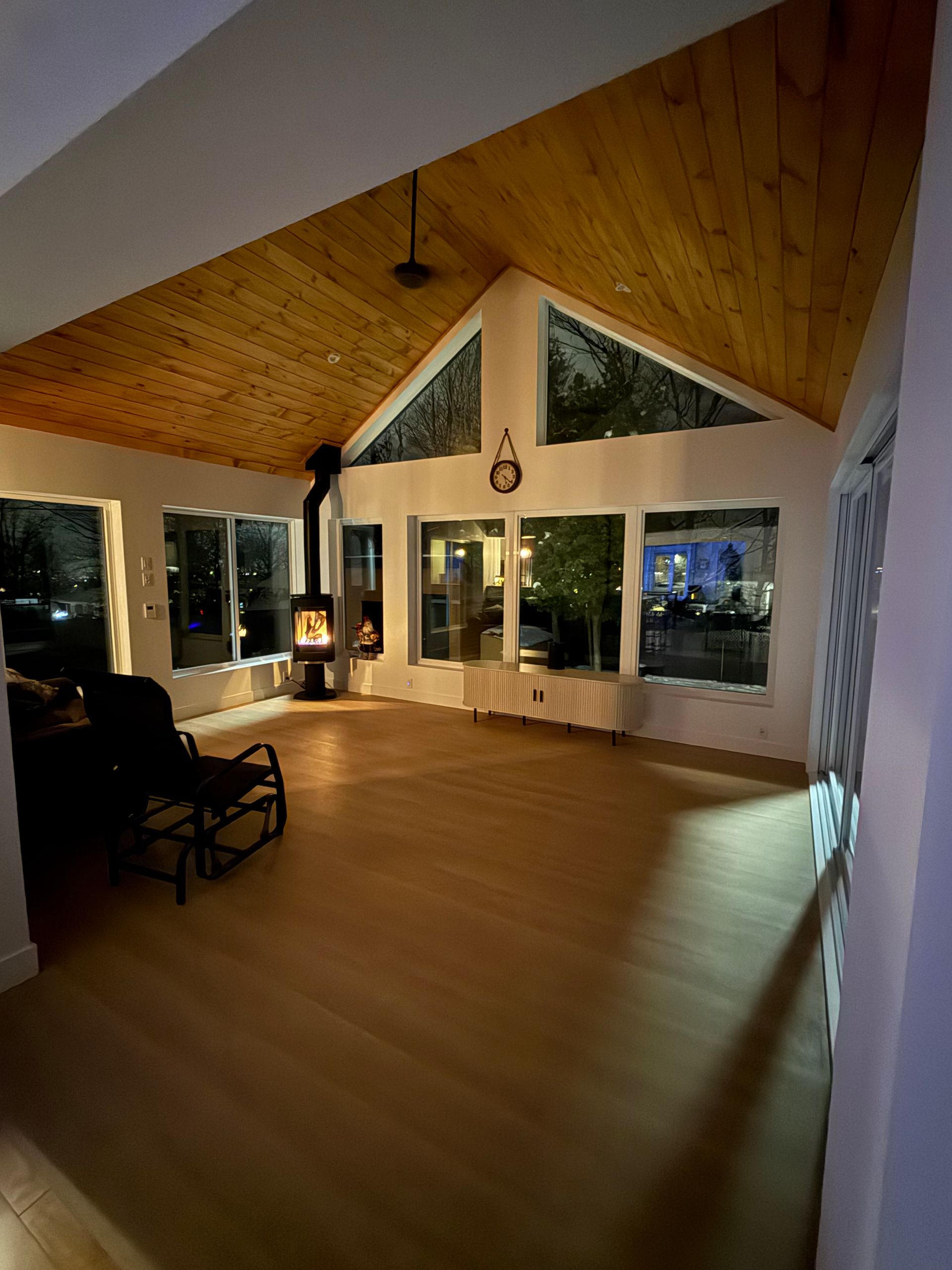 Intérieur chaleureux : parquet et plafond en bois clair, grandes fenêtres, poêle à bois, fauteuil à bascule.