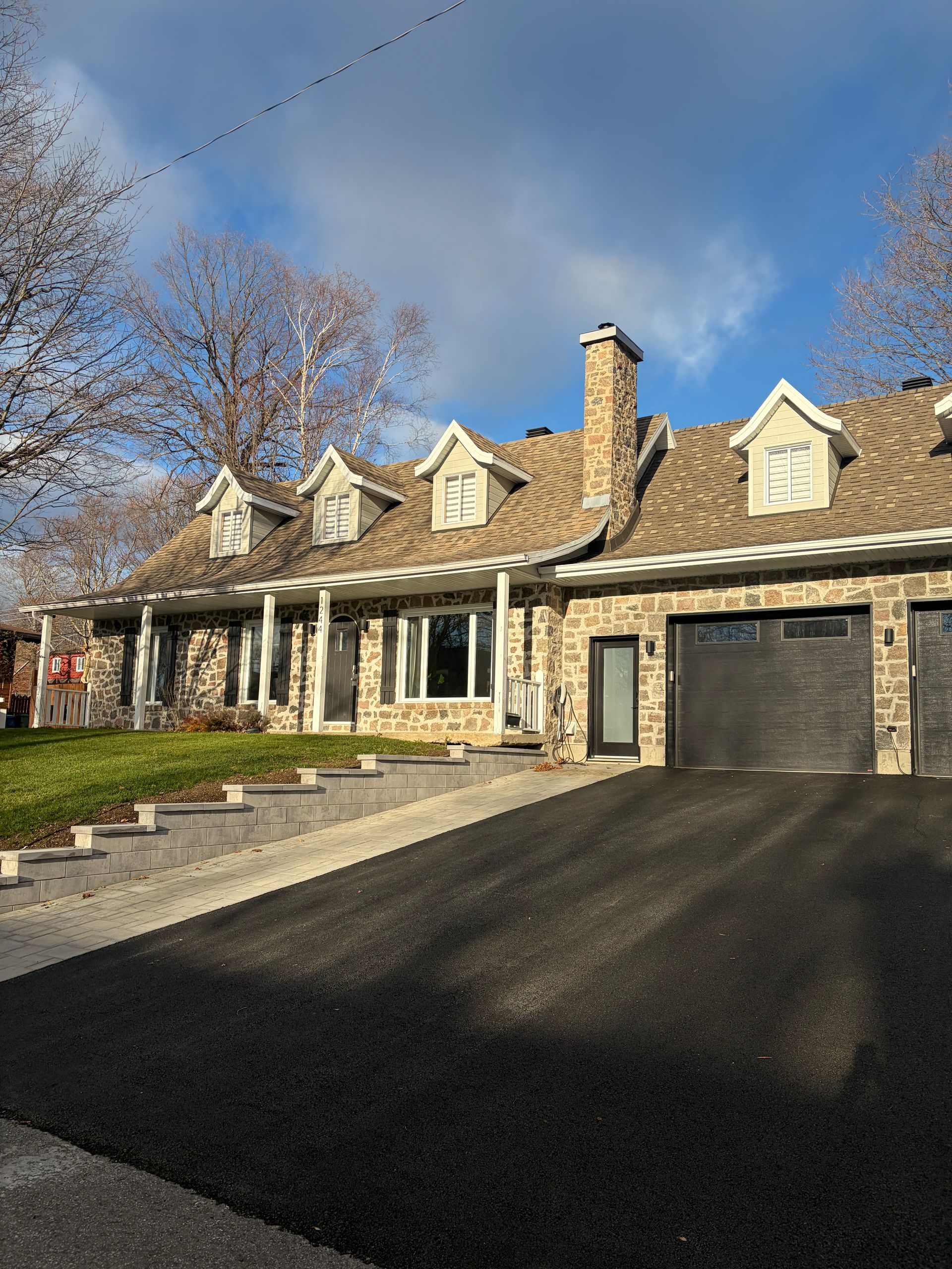 Maison en pierre avec toit à pignons, lucarnes, cheminée et portes de garage noires ; allée asphaltée.