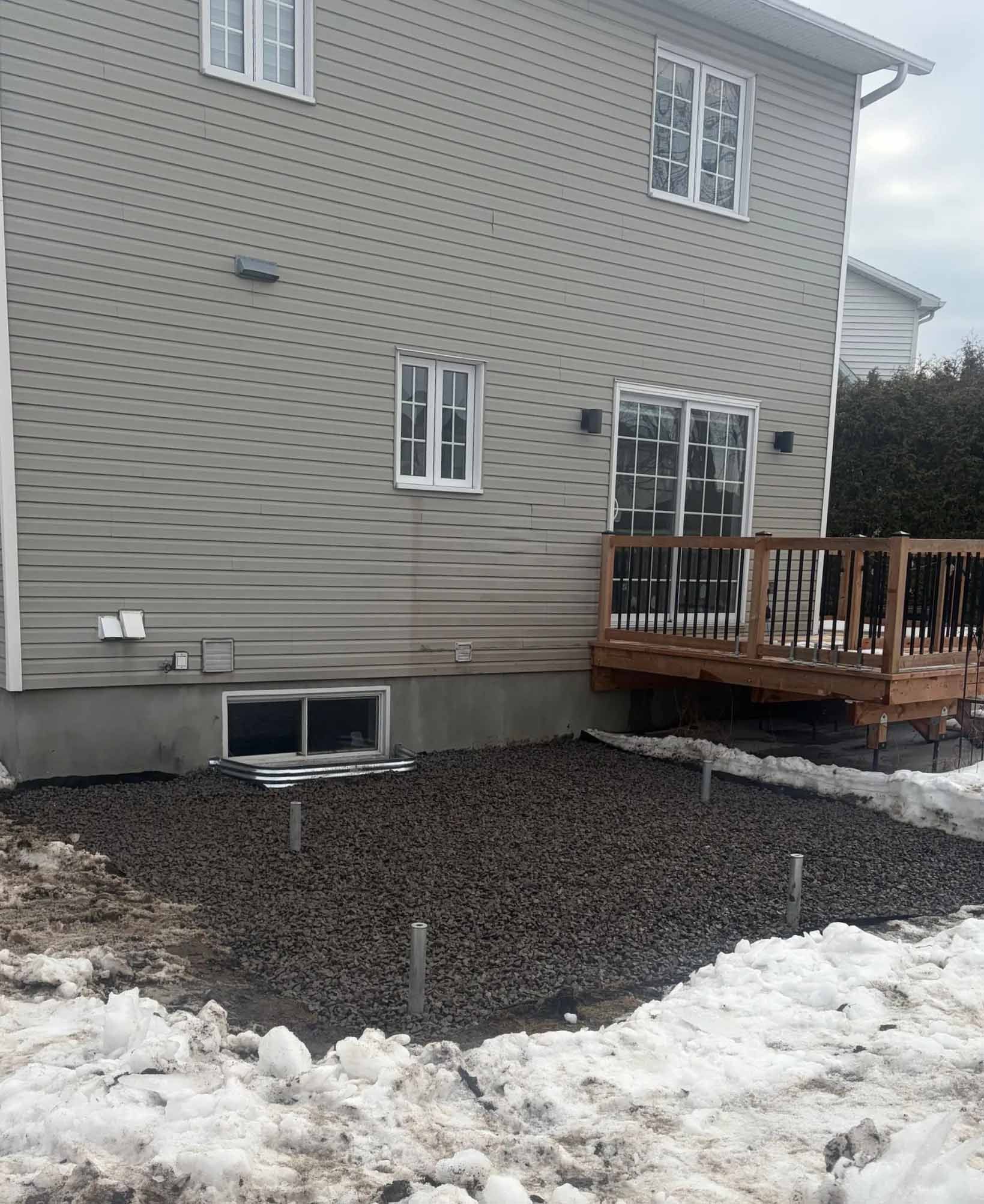 Terrasse arrière jouxtant une maison grise, avec de la neige par endroits sur un sol de gravier sombre.