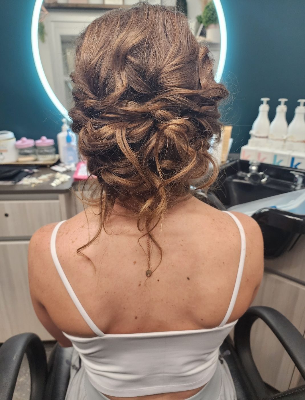 Woman with updo hairstyle, wearing a white top, seated in a salon chair.