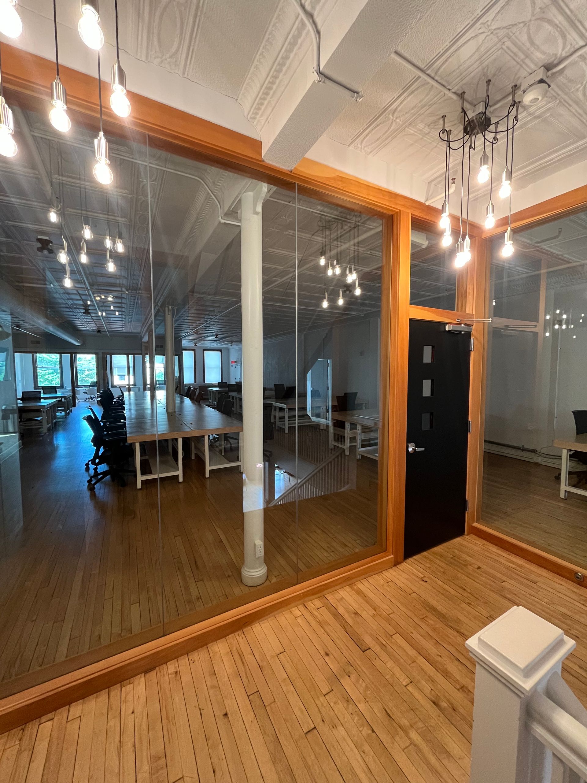 Glass-walled office interior with wood floors, white columns, and pendant lights reflected in the glass.