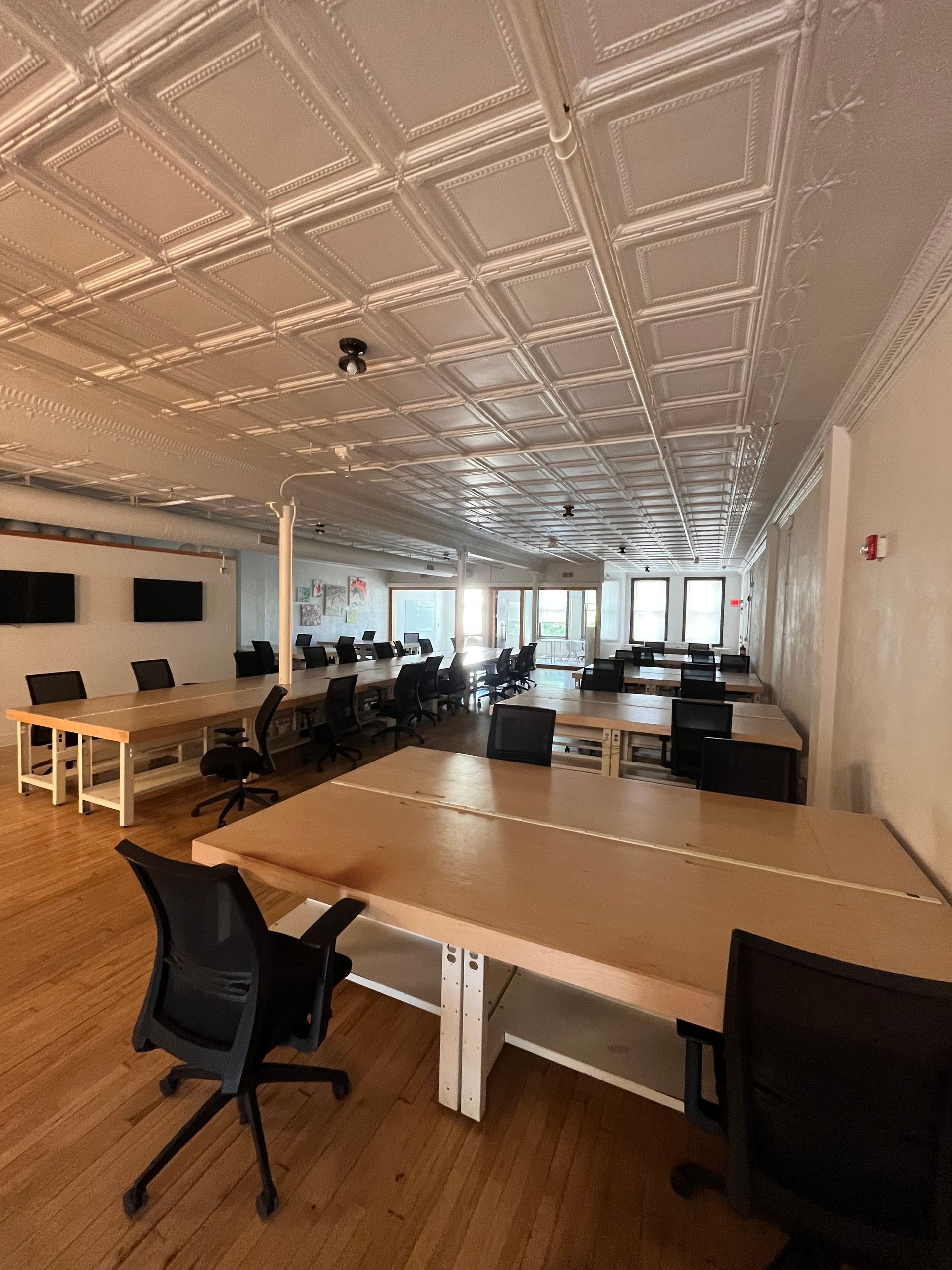 Open office with long desks, black chairs, computers, and large windows at the far end.