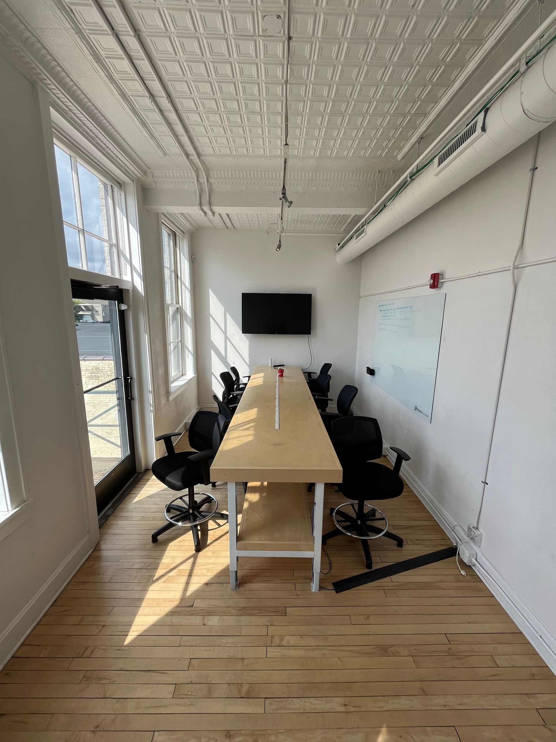 Modern conference room with long table, black chairs, wall-mounted screen, and whiteboard by tall windows