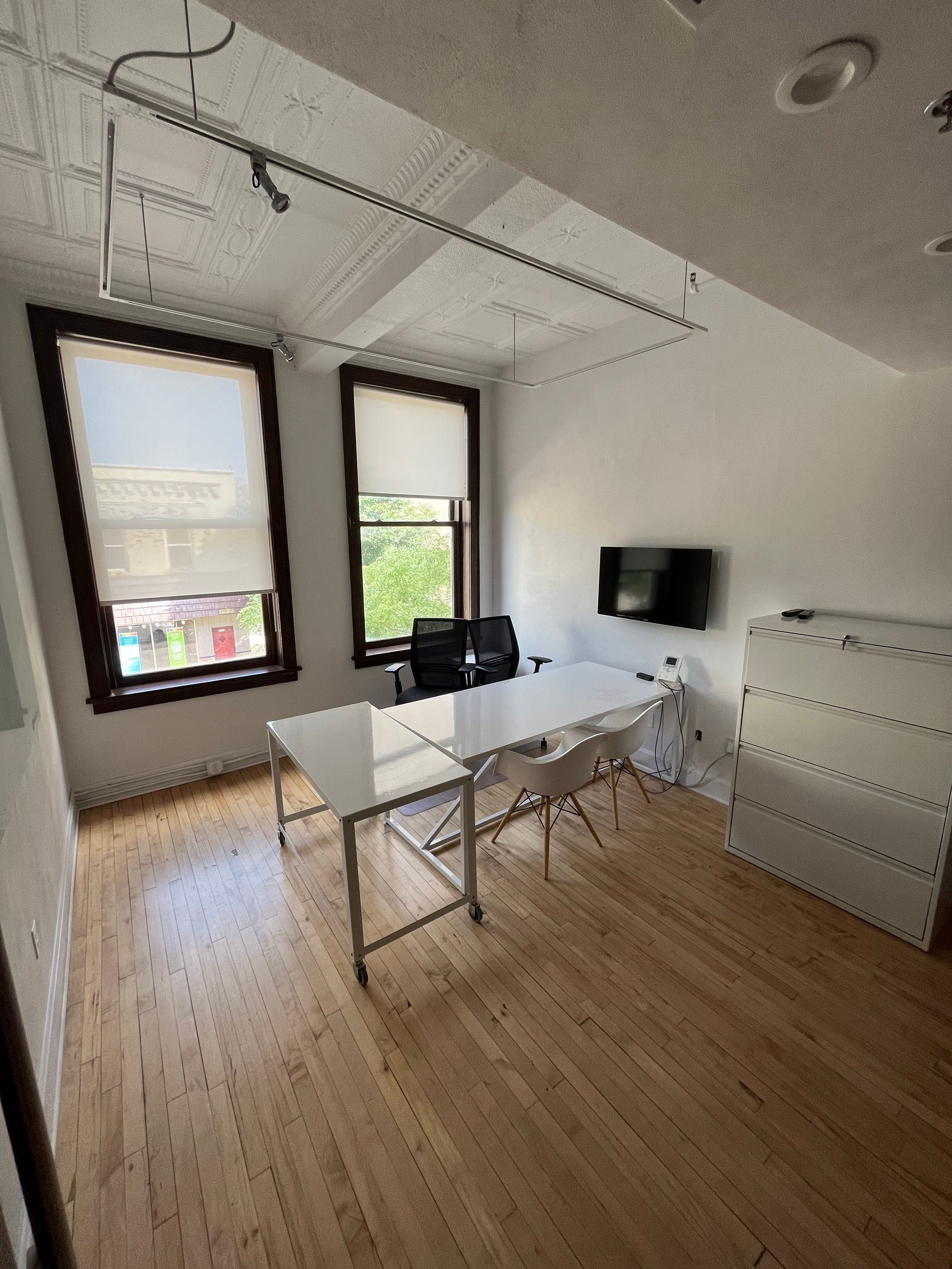 Bright home office with desk, chairs, wall-mounted TV, white cabinets, and large windows.