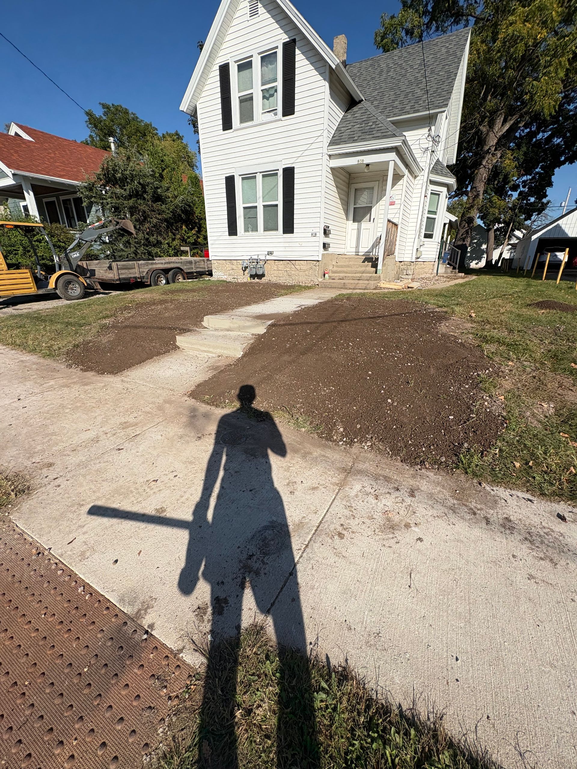 White two-story house with a stone walkway and bare landscaping, viewed from a driveway with a long shadow in front