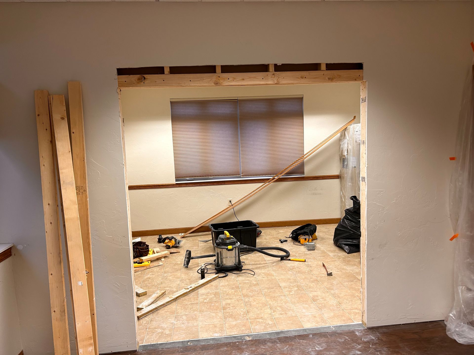 Partially demolished interior wall opening to a messy room with tools, debris, and a window with blinds